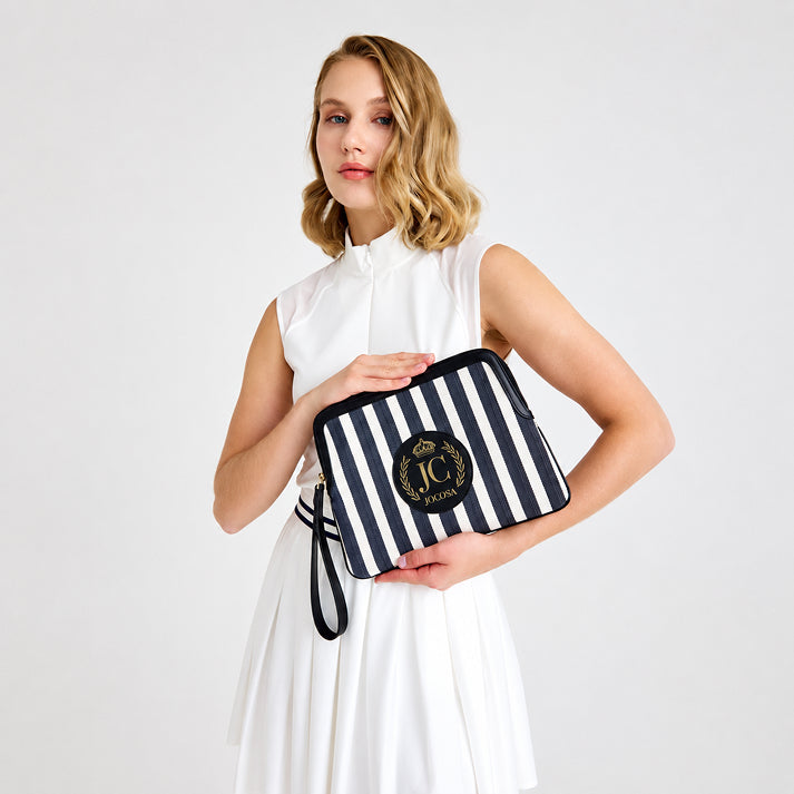 Female model wearing a white tennis outfit, holding the JOCOSA Locker Pouch in ivory and navy striped canvas with black leather trim, a wrist strap and leather logo with gold foil detailing.
