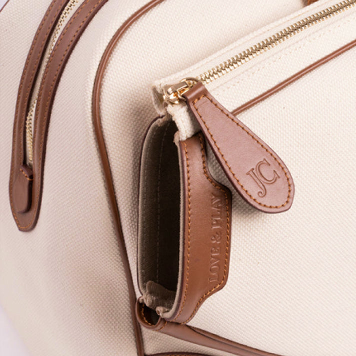 Close-up of the JOCOSA Club Bag's tennis racket pocket opening in ivory canvas with camel leather trim, gold-tone zipper and embossed leather zipper pull.