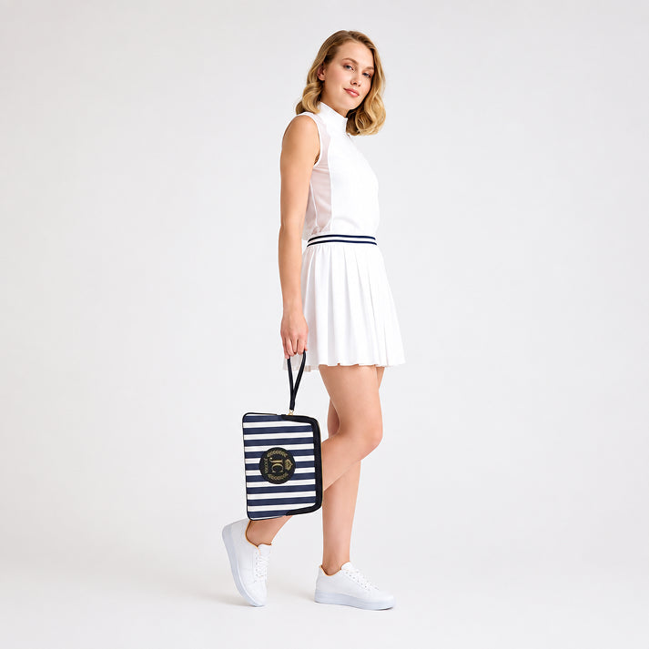 Female model wearing a white tennis outfit and white sneakers, carrying the JOCOSA Locker Pouch by its wrist strap in ivory and navy striped canvas with black leather trim and leather logo with gold foil detailing.