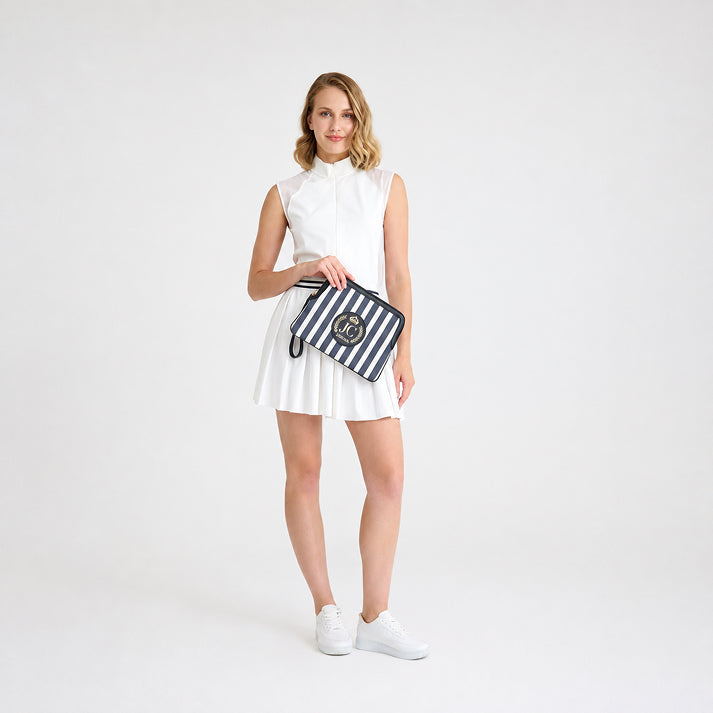 Full-length view of a female model wearing a white tennis outfit and white sneakers, holding the JOCOSA Locker Pouch in ivory and navy striped canvas with black leather trim, a wrist strap and leather logo with gold foil detailing.