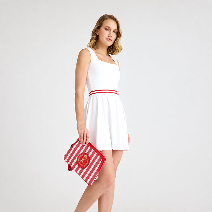 Female model wearing a white tennis outfit, holding the JOCOSA Locker Pouch in ivory and red striped canvas with red leather trim, a wrist strap and leather logo with gold foil detailing.