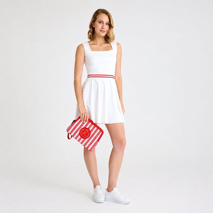 Full-length view of a female model wearing a white tennis outfit and white sneakers, holding the JOCOSA Locker Pouch in ivory and red striped canvas with red leather trim, a wrist strap and leather logo with gold foil detailing.