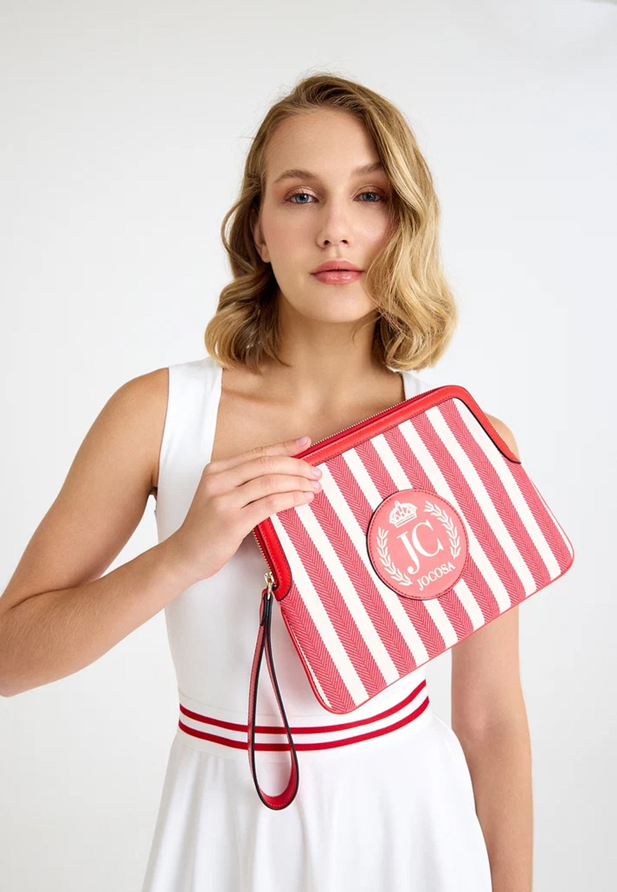 Female model wearing a white tennis outfit, holding the JOCOSA Locker Pouch in ivory and red striped canvas with red leather trim, a wrist strap and leather logo with gold foil detailing.