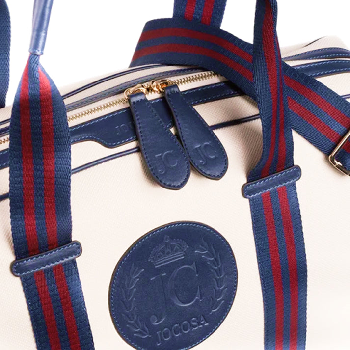 Close-up of the JOCOSA Club Carryall in ivory canvas with navy leather trim, navy and bordeaux striped handles, gold-tone zippers, embossed leather zipper pulls and embossed leather logo.